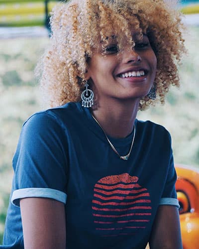 black woman with blond hair wearing purple tshirt with sunset