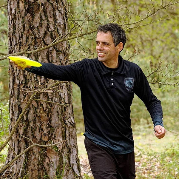 man wearing a black long sleved polo shirt playing with a yellow disc golf in the woods