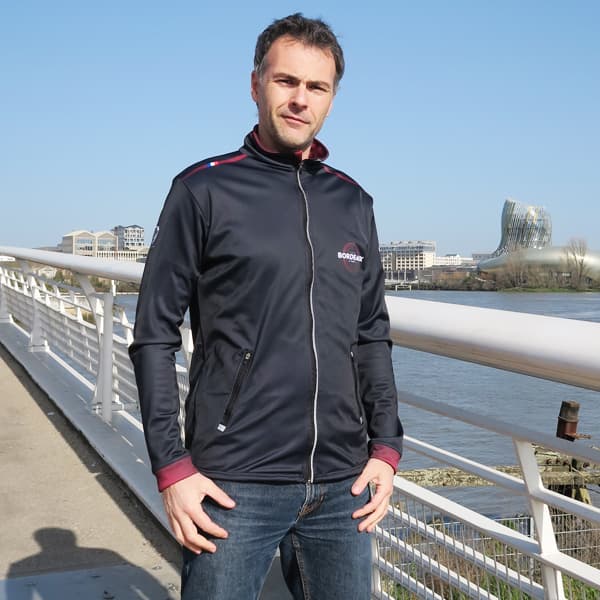 man standing on a bridge wearing a black sport jacket with the cité du vin in the background