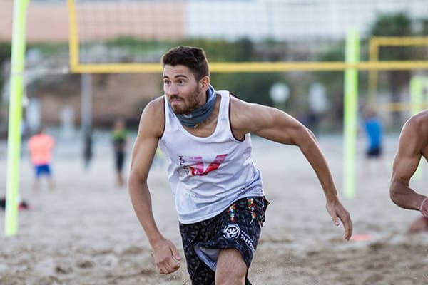 man running on beach wearing a white tank top with volleyball net in the background